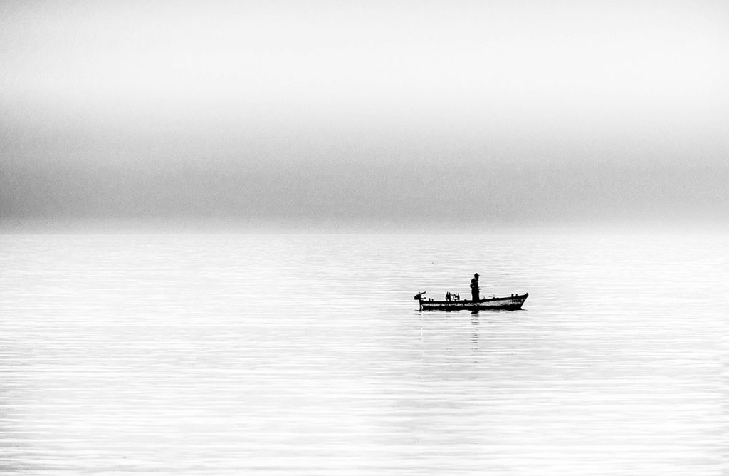 Black-and-white landscape photo of a lone fisherman at sunset