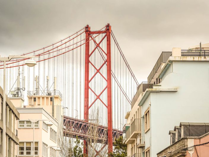 Cityscape photo in Lisbon of the bridge crossing the Tagus River. The photo shows an urban street scene with a large, red suspension bridge rising above the buildings in the background. The buildings in the foreground are low-rise with light pastel colours, while the bridge attracts attention with its robust metal construction and red colour. The sky is overcast and threatening, adding a grey hue to the scene. The mood of the photo is an interesting mix of urban structure and industrial dominance. The contrasts between the soft colour palette of the buildings and the bright red bridge give the photo visual layering. The vertical lines of the buildings and the bridge create a sense of stability and structure, while the heavy cloud sky hangs over everything and adds a certain tension to the image. The photo seems to speak to the relationship between urban development and industrial power, and how these two elements interact.