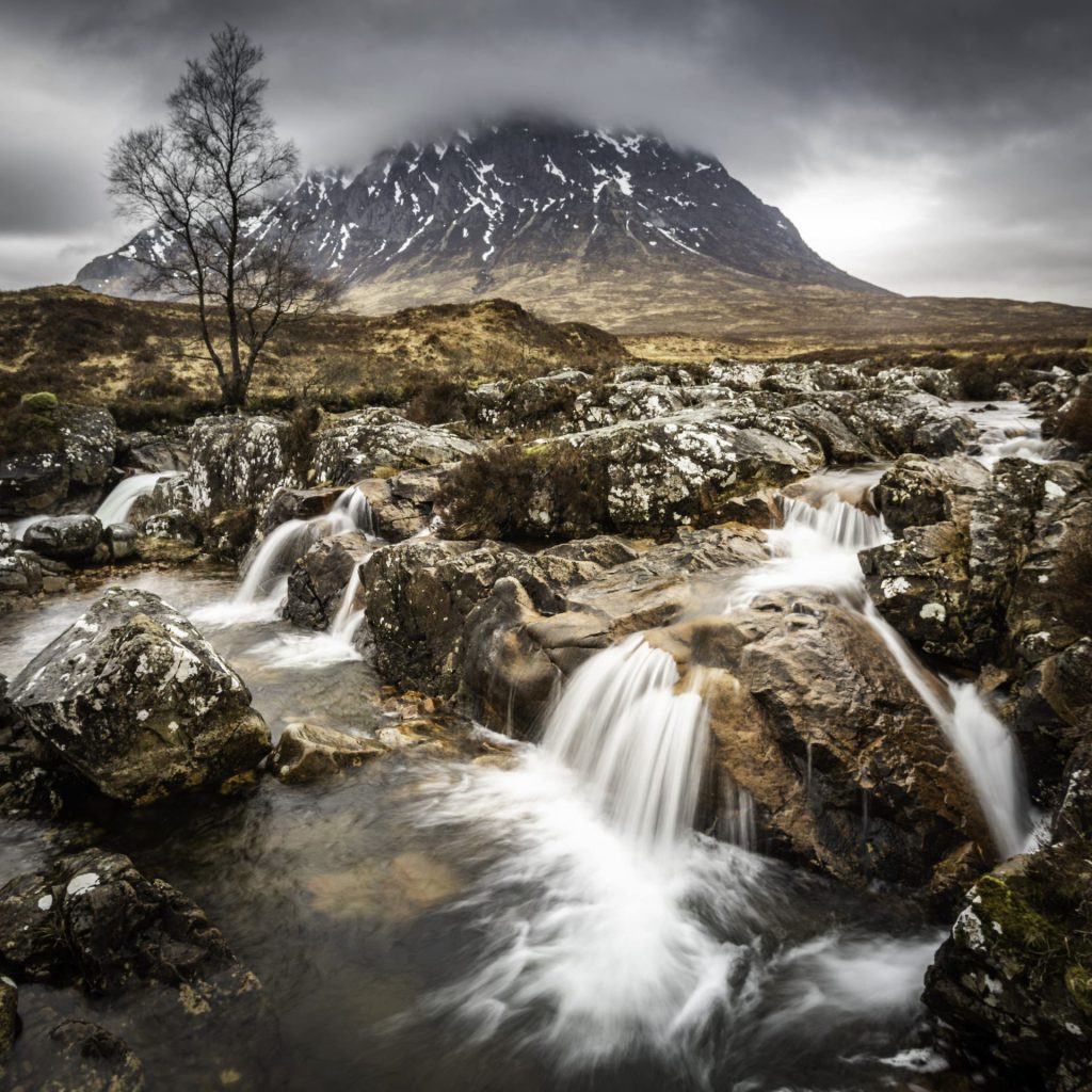 "Highland Pulse" vividly captures the raw and untamed beauty of the Scottish Highlands (Glencoe). The foreground is dominated by a rugged, dynamic stream, tumbling over rocks in a series of miniature waterfalls that create a lively, frothy spectacle. The soft, flowing water is beautifully rendered with a long exposure, giving it a ghostly smoothness that contrasts with the harsh, jagged rocks. In the background, the majestic mountain stands stoic and imposing under a moody, cloud-filled sky, adding a sense of enduring grandeur to the scene. The landscape's muted colours, dominated by browns and greys, evoke the chill of a typical highland atmosphere. The composition is balanced, with the eye being naturally drawn from the turbulent waters in the foreground up to the mountain peak in the distance. The choice to capture the movement of water with a long exposure adds an element of serenity and a magical quality, contrasting with the static and rugged mountain.