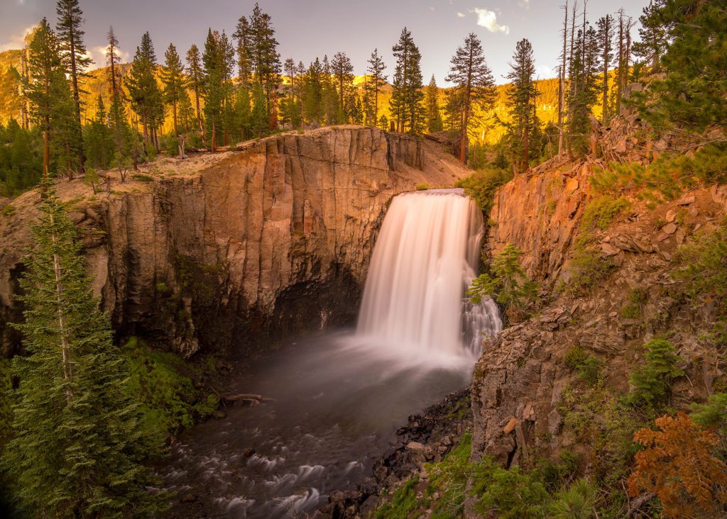 "Amber Cascade Serenity", captured during the golden hour, this photograph is a symphony of nature's majesty and tranquillity. The central waterfall, a dynamic burst of white, plummets into the heart of the image with a soft ferocity framed by the rigid grandeur of the cliffs. The subdued light bathes the scene in warm amber tones, lending the trees a fiery aura contrasting with the fall's cool mist. The long exposure creates a silken texture in the water, emphasising movement amidst the stillness of the surrounding forest and rock.
