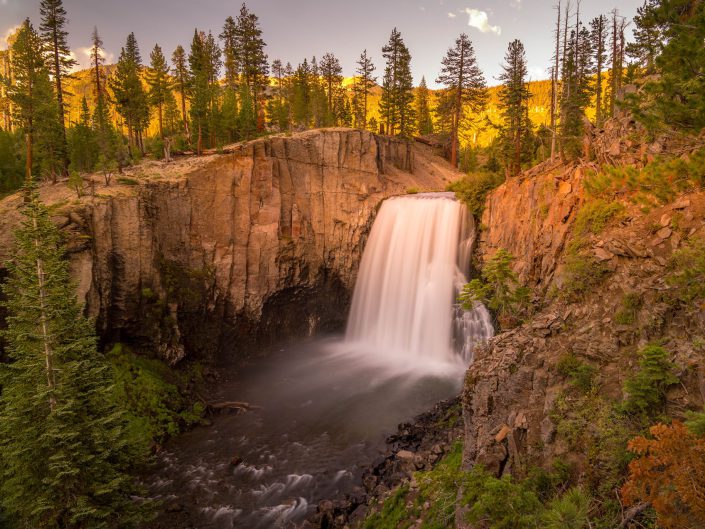 "Amber Cascade Serenity", captured during the golden hour, this photograph is a symphony of nature's majesty and tranquillity. The central waterfall, a dynamic burst of white, plummets into the heart of the image with a soft ferocity framed by the rigid grandeur of the cliffs. The subdued light bathes the scene in warm amber tones, lending the trees a fiery aura contrasting with the fall's cool mist. The long exposure creates a silken texture in the water, emphasising movement amidst the stillness of the surrounding forest and rock. 