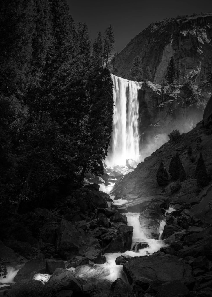"Monochrome Majesty" is a stunning black-and-white photograph that captures Vernal Falls's raw power and elegance. The absence of colour focuses the viewer's attention on the textures and contrasts within the scene—from the misty spray of the falls to the rugged, angular boulders below. Towering conifers stand sentinel on either side, their dark silhouettes framing this display of nature's force. The play of light and shadow, alongside the cascading water, creates a dynamic yet timeless image that encapsulates the essence of the wild. This piece is an example of landscape photography that uses monochromatic tones to enhance the subject's natural drama and beauty. The composition is meticulous, and the choice to omit colour magnifies the impact of the natural forms and the intensity of the water's flow.