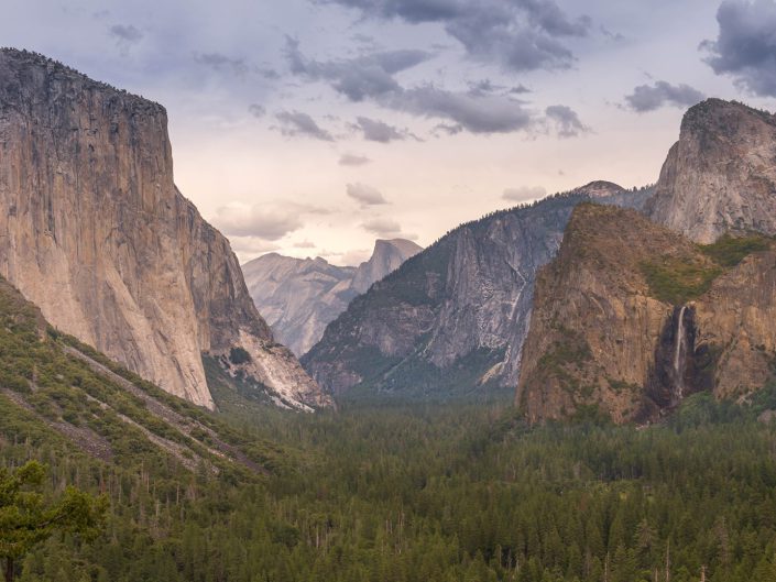 "Majesty of the Valley": This photo showcases the breathtaking Yosemite Valley, captured with a wide lens that embraces its expansive grandeur. The left side of the frame is dominated by El Capitan, a monumental granite monolith that rises starkly against the sky. Its sheer cliff face is a testament to geological time and the forces of nature. To the right, Bridal veil Fall gracefully plummets, its mist catching the light and adding a sense of movement to the stillness of the cliffs. In the distance, the iconic Half Dome can be seen, its distinct shape recognisable even in silhouette. The valley floor is carpeted with a dense forest of pine trees, and their deep greens provide a rich base to the towering cliffs. Overhead, a dramatic sky with clouds tinged by the sun's soft glow because of the late afternoon light offers a dynamic and changing atmosphere to the scene. In this photograph, the choice was not to focus on the waterfall centrally, but it is still an integral part of this expansive view. 