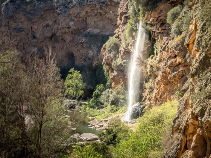 "Whispering Cascade" presents a serene and almost otherworldly depiction of nature's elegance. Centred in the image, the waterfall pours its silky waters from a height framed by abundant greenery. The light filtering through the canopy above lends a soft glow to the water and illuminates the leaves, enhancing the overall ethereal quality of the scene. The slow shutter speed technique here allows the water to appear as a continuous, smooth veil, contrasting beautifully with the crisp texture of the surrounding vegetation. The composition, while slightly central, effectively draws the viewer's eye straight to the waterfall, making it the undeniable focal point of the piece. 