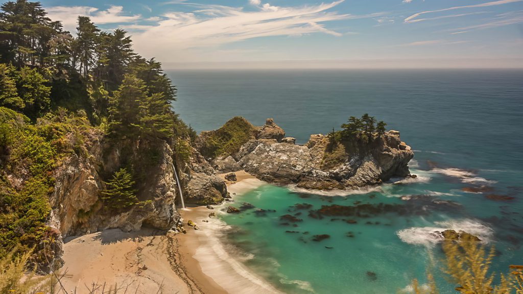 "Coastal Reverie" is a breathtaking photograph that captures the magnificence of McWay Falls, located at Julia Pfeiffer Burns State Park, nestled along the picturesque Big Sur coastline in California. The falls cascade gracefully onto a secluded beach, creating a thin mist that merges with the turquoise waters of the Pacific Ocean. The cove is encircled by rugged cliffs and adorned with lush greenery, where the trees tenaciously cling to rocky outcrops, defying the salty sea breeze. The expanse of the ocean stretches to the horizon, its surface shimmering under a sky dotted with wispy clouds, hinting at a serene and sunny day. The clear, aquamarine hue of the protected bay contrasts sharply with the deep blue of the open sea, highlighting the peaceful isolation of this hidden gem. Though a bright sunny day may not be the ideal time for a good landscape photo, this picture successfully captures a magical moment in a place where the elements of land, sea, and sky harmoniously converge, making it an ideal destination for nature lovers and photography enthusiasts.