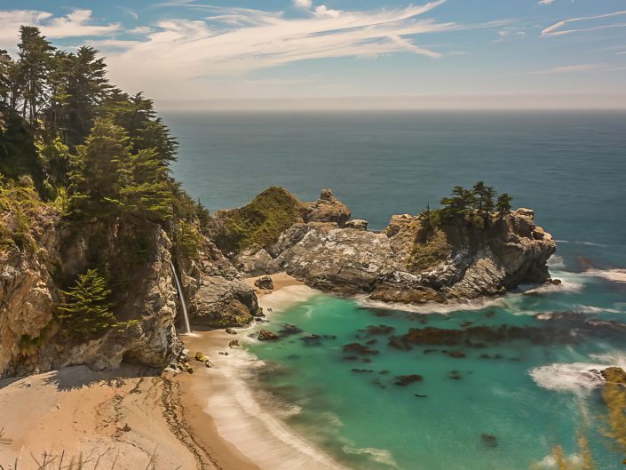 "Coastal Reverie" is a breathtaking photograph that captures the magnificence of McWay Falls, located at Julia Pfeiffer Burns State Park, nestled along the picturesque Big Sur coastline in California. The falls cascade gracefully onto a secluded beach, creating a thin mist that merges with the turquoise waters of the Pacific Ocean. The cove is encircled by rugged cliffs and adorned with lush greenery, where the trees tenaciously cling to rocky outcrops, defying the salty sea breeze. The expanse of the ocean stretches to the horizon, its surface shimmering under a sky dotted with wispy clouds, hinting at a serene and sunny day. The clear, aquamarine hue of the protected bay contrasts sharply with the deep blue of the open sea, highlighting the peaceful isolation of this hidden gem. Though a bright sunny day may not be the ideal time for a good landscape photo, this picture successfully captures a magical moment in a place where the elements of land, sea, and sky harmoniously converge, making it an ideal destination for nature lovers and photography enthusiasts.