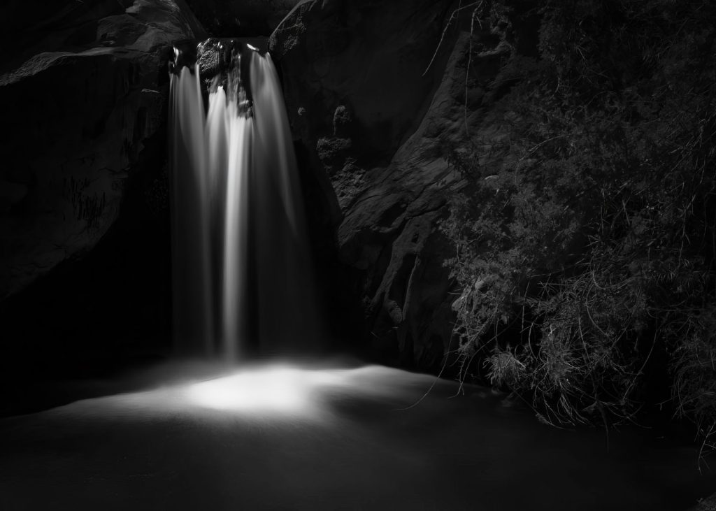 This evocative black-and-white photograph captures the haunting beauty of a small solitary waterfall near Palm Springs in a seemingly nocturnal setting, which wasn’t the case. The waterfall flows smoothly over a cliff, forming a stark white ribbon against the dark, shadowy backdrop. The long exposure in capturing the image creates a silky texture in the water's motion, which pools into a glowing basin of light at the foot of the falls. The surrounding rocks and vegetation are shrouded in darkness, with just enough detail to suggest a rugged and untamed landscape. This minimalist approach draws all the attention to the luminous cascade, emphasising the interplay of light and darkness and the contrast between the fluidity of the water and the solidity of the rock. The choice of black and white enhances the timeless quality of the scene and focuses the viewer on the textures and light rather than the distractions of colour.