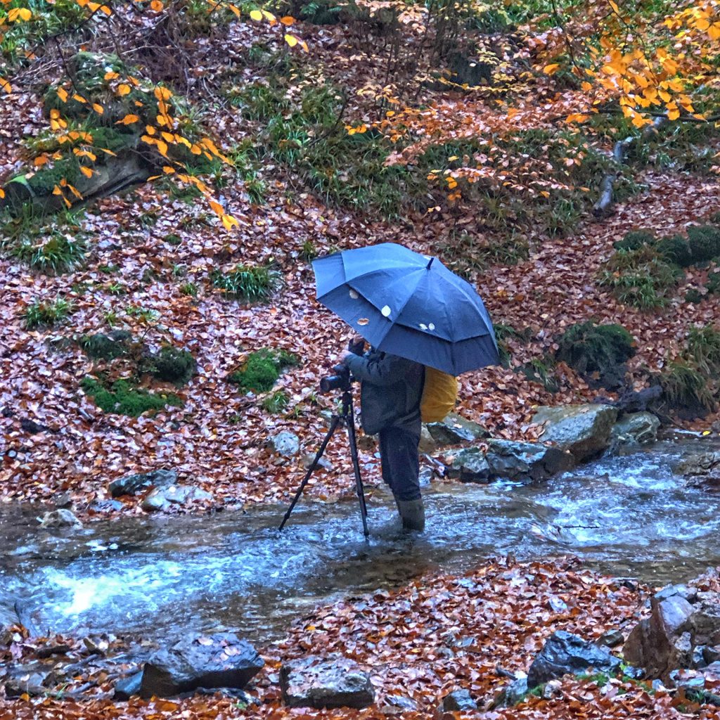 The photo shows me engaged in outdoor landscape photography, standing in a shallow stream sheltered by a dark umbrella and wearing rubber boots and raincoats. The tripod is set firmly in the water, capturing the movement of the stream, a small waterfall not visible in this photo.