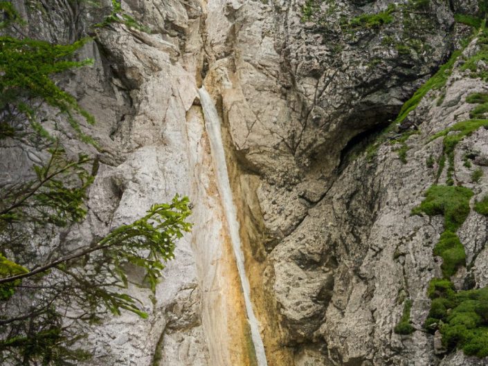 The image depicts a breathtaking natural waterfall surging through a rugged cliff. The waterfall is tall, slender, and robust, forming a striking contrast against the weathered grey-white stone. The surrounding rock formations are jagged and imposing, with patches of vegetation clinging to their surfaces to soften their rough appearance. At the waterfall's base, a small group of individuals provides a sense of scale, accentuating the grandeur of the waterfall. This inclusion adds a relatable aspect and an adventurous atmosphere, inviting viewers to imagine the sound of the water and the experience of witnessing such a spectacle in person. 