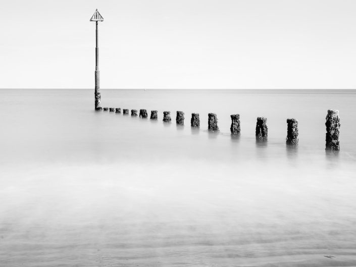 The picture shows a minimalist coastal landscape, with a row of wooden poles leading into the sea. The posts are covered with algae and mussels, highlighting their weathered and rough texture. At the end of the row is a marker pole with a triangular sign on top. The sea has been smoothed by a long exposure, giving the water's surface an ethereal, misty effect. The photo exudes a sense of stillness and infinity. Using black and white enhances the abstraction and focuses attention on the shapes and lines in the image rather than on colour. The emptiness of the horizon and the symmetrical repetition of the poles contribute to a sense of tranquillity and timelessness. The composition uses guiding lines. The row of poles draws the viewer's gaze from the foreground to the horizon, adding depth and direction to the image. The vertical lines of the poles contrast with the horizontal lines of the sea and horizon. The repetition of the poles creates a rhythm and structure in the composition. The photo has a strong balance thanks to the symmetrical placement of the poles, which act as a natural anchor point against the soft, diffuse background. The diffused light creates a smooth, even exposure without harsh shadows. The long exposure time has softened the water, creating a calm and serene atmosphere.