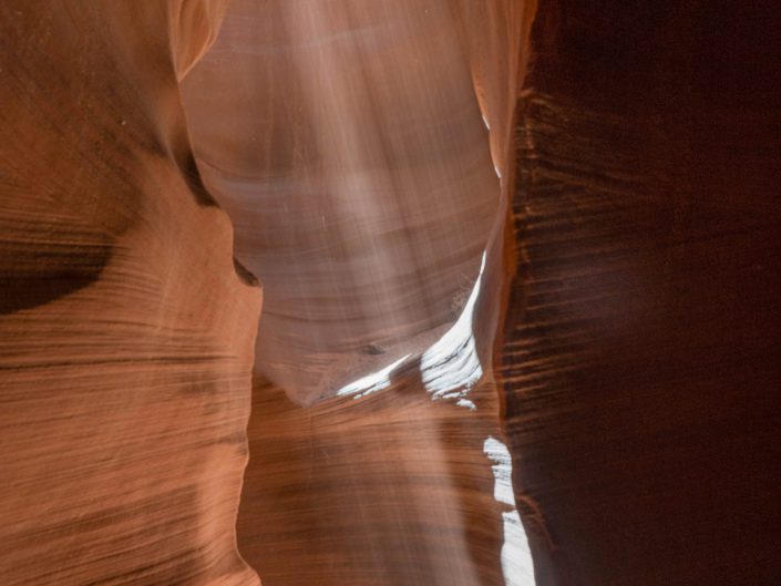 The photo shows a narrow, vertical canyon in Antelope Canyon, through which a strong beam of natural light shines. The canyon walls are smooth and eroded by time, with deep shadows contrasting with the bright beam of light. The colours of the rocks range from warm orange tones to deep, dark shadows. The photo captures a magical moment where the light falls through the canyon's narrow opening, creating an almost spiritual atmosphere. The beam of light draws attention and highlights the organic shapes of the rocks, which fall along the walls like a soft veil. The combination of light and shadow, along with the warm colours of the rock, gives the picture a dramatic yet serene feel. The image symbolises the power of light and its ability to reach even the darkest places, giving it a contemplative and almost mystical quality.