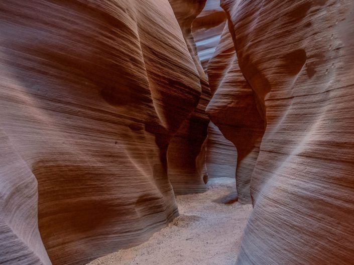 The picture shows a narrow passage in a slot canyon. The walls have been eroded by years of erosion, resulting in smooth, undulating shapes. The colours of the rock walls range from deep, warm hues of orange and red to cooler shades of blue and purple. Covered with fine sand, the photograph exudes an air of mystery and serenity. The narrow passage invites the viewer to wonder where the path leads, evoking a sense of adventure and discovery. The lighting is soft, with the light falling diffusely through the narrow opening at the top, subtly highlighting the textures and colours of the rocks. The photograph balances between abstraction and realism, focusing on the shapes and the play of light and shadow, contributing to a sense of timelessness.