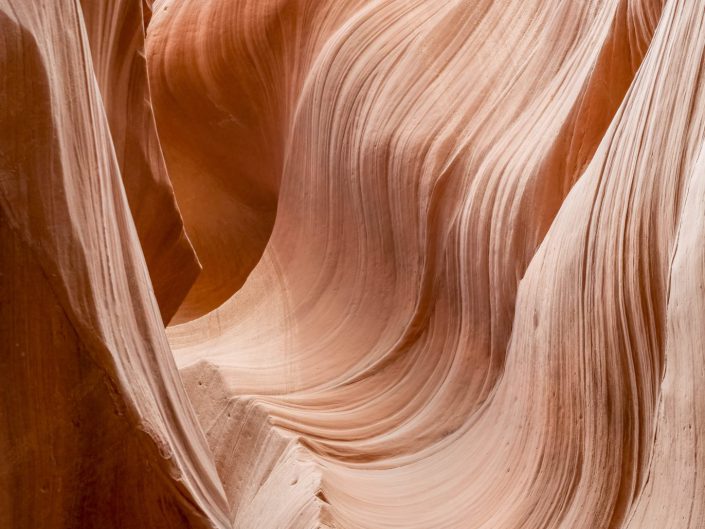 The photo shows a close-up of sandstone walls eroded by water and wind, resulting in flowing, organic shapes and textures. The colours range from soft pinks to deep orange tones, highlighting the natural beauty of the rock, and the picture exudes a serene and timeless atmosphere, the result of thousands of years of erosion. The gentle curves and lines in the rocks suggest movement and harmony, as if the canyon walls were dancing with the light. The light falls in diffusely, enhancing the subtle textures and nuances of colour. The photograph captures a moment in nature that is both powerful and elegant, with an almost mystical quality that invites the viewer to stand still and admire.