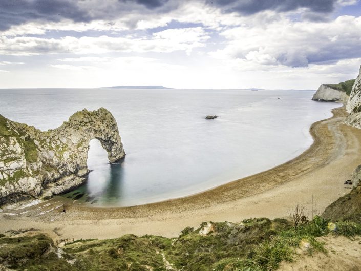 The picture shows an iconic coastal scene, with Durdle Door at its centre. Durdle Door is a natural limestone arch on the Jurassic Coast in Dorset, England. The arch rises majestically from the sea, framed by a tranquil beach and dramatic cliffs. The composition captures a moment of tranquillity, where the gentle water kisses the shore and the overcast sky embraces the landscape.