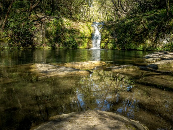 The picture shows a small waterfall flowing into a calm, shallow pool in the forest. The atmosphere is serene and soothing, enhanced by the soft light falling through the trees and the calm water reflecting the surroundings. The image exudes a sense of peace and balance, as if nature is in perfect harmony now. The artistic qualities of the picture lie in its composition and the subtle balance between light and shadow, which adds to its contemplative mood. 
The photograph's composition is carefully constructed to emphasise symmetry and reflection. The horizontal lines of the stones in the foreground and the water surface create a base that leads the viewer to the central waterfall. The vertical line of the waterfall, which dominates the centre of the image, provides a natural focal point. The trees and greenery hug the image, creating a natural frame that guides the viewer's eye to the waterfall and the reflection in the water. There is a clear balance between the elements in the picture; the reflection in the water reflects the aforementioned elements, adding a sense of rhythm and harmony. The techniques used, such as the lighting (diffused light that reveals the details of the greenery and water), the focus on the waterfall and the reflection, enhance the serene atmosphere.