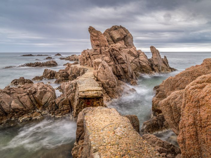 The photograph shows a rugged coastal landscape with impressive rock formations rising from the sea. In the foreground, a narrow path of stone can be seen meandering through the rocks, seemingly disappearing into the distance, towards a large, striking rock formation that forms the centrepiece of the composition. The sea surrounds the rocks and creates a misty effect due to the long exposure time, contributing to an atmosphere of mystery and timelessness. The sky is overcast and heavy, with shades of grey and blue giving the landscape a cool, serene look. The photograph has a strong sense of depth and direction, partly due to the path that leads the viewer through the landscape to the dramatic climax of the composition: the largest rock formation. The balance between the textures of the rocks and the smooth, almost ethereal water captured by the long exposure gives the photo a dynamic contrast between solidity and movement. This contrast is further enhanced by the natural colour tones of the rocks, ranging from warm, earthy tones to cooler, stony colours, and the subtle blues of the water and sky.