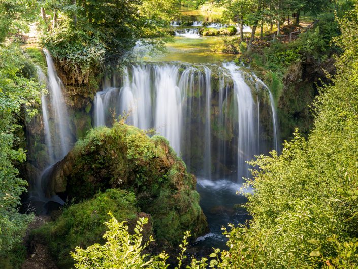 "Eternal Flow". This picture shows a breathtaking waterfall surrounded by lush green vegetation. The water falls gracefully into a naturally formed basin, with the fine mist creating an ethereal atmosphere. The reflection of light on the water's surface and the gentle flow of the water suggest a moment of timeless tranquillity. This shot not only illustrates the beauty of nature, but also gives the viewer a sense of peace and serenity. The composition of this photograph has been considered with a central placement of the waterfall forming the visual focal point. Around the waterfall, the different shades of green of the leaves create a natural frame that guides the eye to the centre. There is a balance between the fluid movements of the water and the static elements such as the rocks and trees. The colours are natural and rich, adding depth and realism to the image.