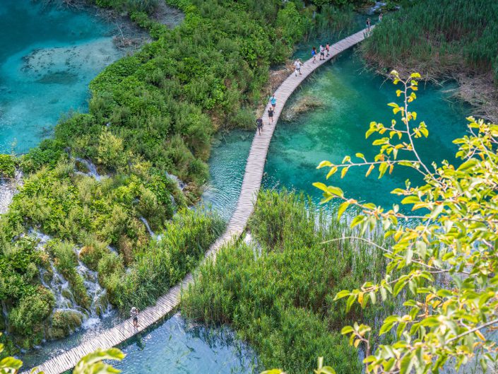 The picture shows a wooden path winding through a beautiful, green-covered environment with clear blue and green waters of the Plitvice Lakes in Croatia. People walking along the path give a sense of scale and perspective. The vegetation is lush, and the water is crystal clear, with turquoise and deep blue nuances. Artistically, the photo captures the harmony between man and nature. The winding path breaks up the wild beauty of the landscape but without disturbing it. The shot from a bird's eye view offers a unique view of the natural beauty and human interaction with it. The colours are vibrant but, at the same time, soothing, evoking a sense of adventure and exploration.