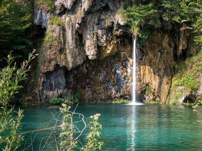 The picture shows an idyllic waterfall flowing from a rocky opening, emptying into a clear, turquoise-coloured lake. Surrounded by lush greenery and rugged rock walls, the scene exudes an atmosphere of tranquillity and pristine nature. The water seems to glisten under the light of the sun, adding to the magical aura of the location. The vertical line of the waterfall contrasts beautifully with the horizontal lines of the water's surface. The rocks offer a rough texture that compliments the soft, smooth surface of the water. The composition is balanced with the waterfall centrally placed, providing a natural focus. The colours are vibrant but natural, with an emphasis on greens and the turquoise colour of the water.