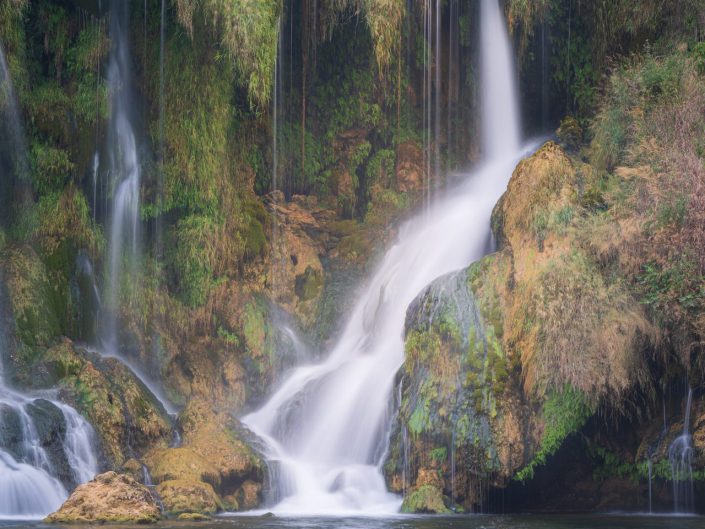 The picture shows a waterfall flowing from a rocky wall into a peaceful water feature. The surroundings are rich in greenery, with moss and ferns covering the rocks, giving the scene a vibrant and natural look. The water appears almost silky due to the long shutter speed, contributing to a calm and serene atmosphere.

Artistically, the photo highlights the harmony between the powerful movement of the water and the quiet, almost static presence of the vegetation. The soft lighting and saturated colours create a dreamy, ethereal quality that invites the viewer to linger longer and immerse themselves in the serenity of nature.