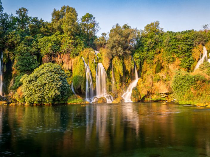 This photo of the Kravica waterfalls in Bosnia shows a beautiful combination of strength and peace, characterised by the powerful water quietly crashing into a calm pool. The greenery surrounding the falls contributes to a sense of fertility and vitality. The light and composition enhance the natural beauty of the location. The vertical lines of the falling water contrast with the horizontal lines of the waterhole and surrounding soil. The natural curves of the vegetation and rocks add softness to the composition. There is a harmonious balance between the elements of water, earth and sky. The lighting is soft due to the late daylight, which enriches the colours of the vegetation and gives the water a sparkling quality.