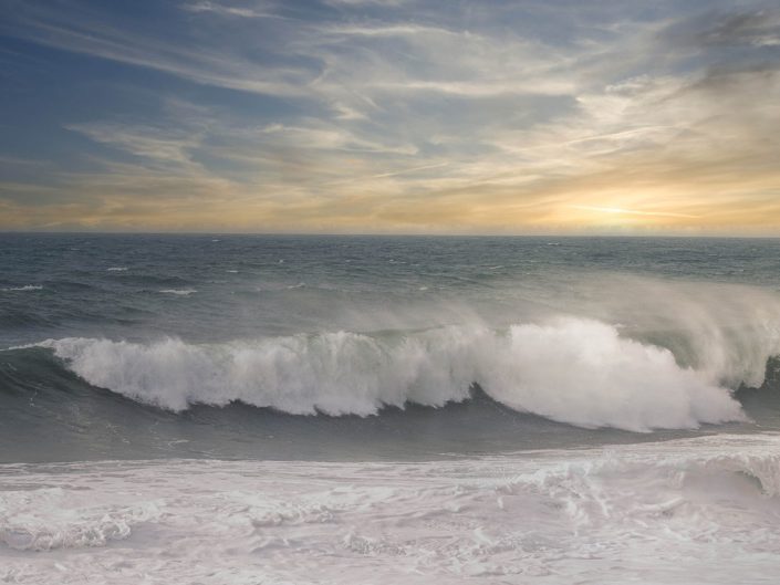 The picture shows a wide expanse of ocean with a dynamic wave about to break on the shore. The sky is partly cloudy, with the sun sending its last rays through the clouds, creating a warm glow just above the horizon. The waves roll powerfully towards the shore, forming white crests.
The mood of the photo exudes calm and strength at the same time. The wave movements and rolling foam seem almost tangible, making the viewer feel connected to nature. The golden hues in the sky contrast nicely with the blue and grey of the ocean, creating a harmonious yet powerful composition. The photograph captures a moment of constant motion and eternal stillness in one image, evoking a deep sense of serenity and infinity.