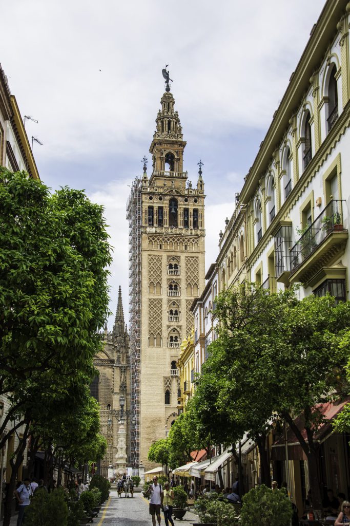 Central to the composition is the Giralda, the tower of Seville Cathedral. The tower is richly detailed, with Moorish motifs in the lower half and Renaissance elements at the top. On both sides of the street are classic Andalusian buildings with balconies and cafes. The street is lined with orange trees. People are walking and a carriage is visible in the background. But the entire tower is in scaffolding.