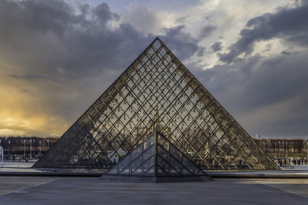 We see a symmetrical composition of the Louvre's famous glass pyramid, photographed straight from the front. The sky is dramatic: yellow-gold and warm on the left, cooler and grey-blue on the right. The glass subtly reflects the light colours. In the foreground, a smaller pyramid repeats the larger one. The sculpture masterfully plays with contrasts: old versus new, light versus dark, simplicity versus complexity. The transparency of the glass makes the structure airy despite its monumentality. The picture has a sacred atmosphere, almost like entering a modern cathedral of knowledge.