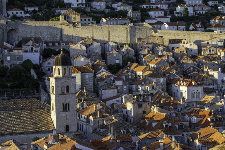 The image shows a classic Dubrovnik (Croatia), with a maze of terracotta roofs under a warm evening light. In the foreground is a striking church tower with a dark dome, and an imposing city wall runs in the background. Artistic: The atmosphere is nostalgic and warm, bathed in golden evening light. The stacking of roofs, the stone tones, and the play of light and shadow create a picturesque scene resembling a still life with urban layers. The presence of modern elements does not make it timeless.