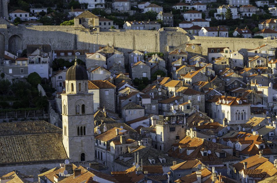 The image shows a classic Dubrovnik (Croatia), with a maze of terracotta roofs under a warm evening light. In the foreground is a striking church tower with a dark dome, and an imposing city wall runs in the background.

Artistic:
The atmosphere is nostalgic and warm, bathed in golden evening light. The stacking of roofs, the stone tones, and the play of light and shadow create a picturesque scene resembling a still life with urban layers. The presence of modern elements does not make it timeless.