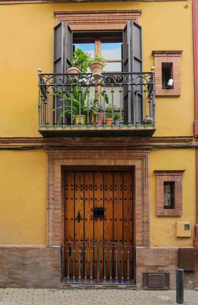 The image shows a front facade of a house with yellow wall, a heavy wooden door with wrought-iron railings below and, above it, a balcony with plants. The balcony has a decorative balustrade and wooden shutters half-opened. Two small windows flank the front door. However the view is littered with cables, security cables and mucht more.
