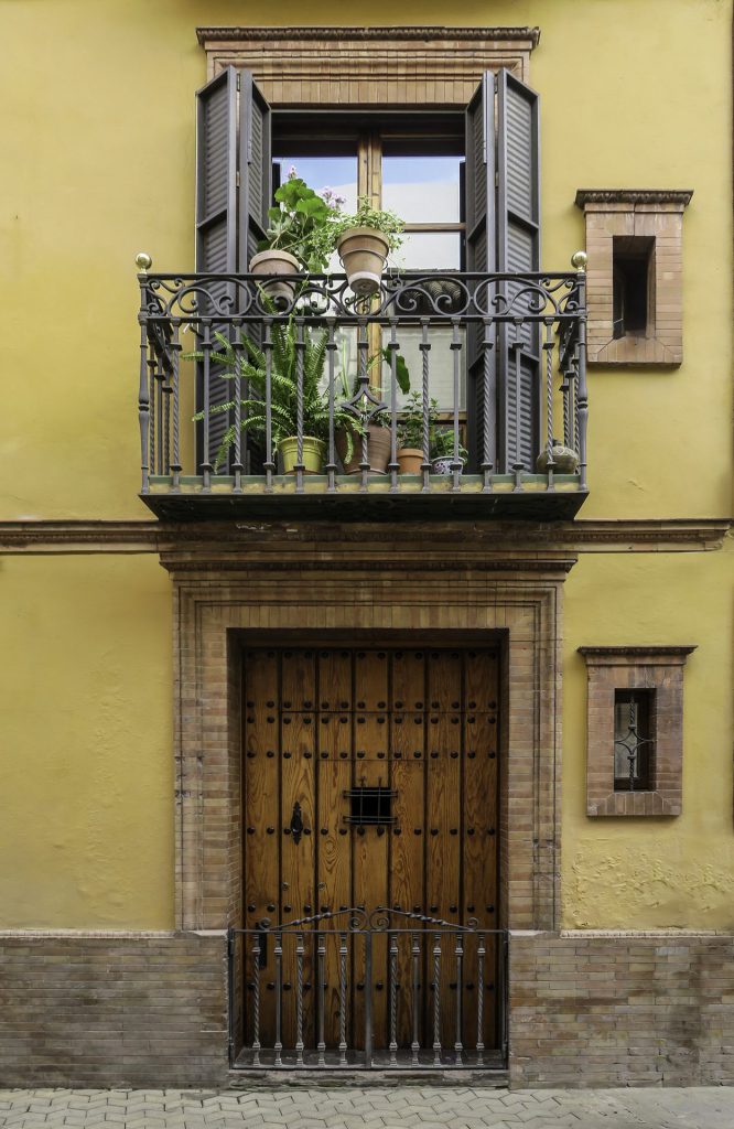 The image shows a front facade of a house with yellow wall, a heavy wooden door with wrought-iron railings below and, above it, a balcony with plants. The balcony has a decorative balustrade and wooden shutters half-opened. Two small windows flank the front door. This picture is a composition of rhythm and tranquillity: repetition in the wrought-iron bars, contrast between warm colours (yellow, brown) and vibrant greenery. The image exudes Mediterranean tranquillity. There is something almost symbolic in how the plants cluster in their balcony mound - a small living stage on a fixed background.