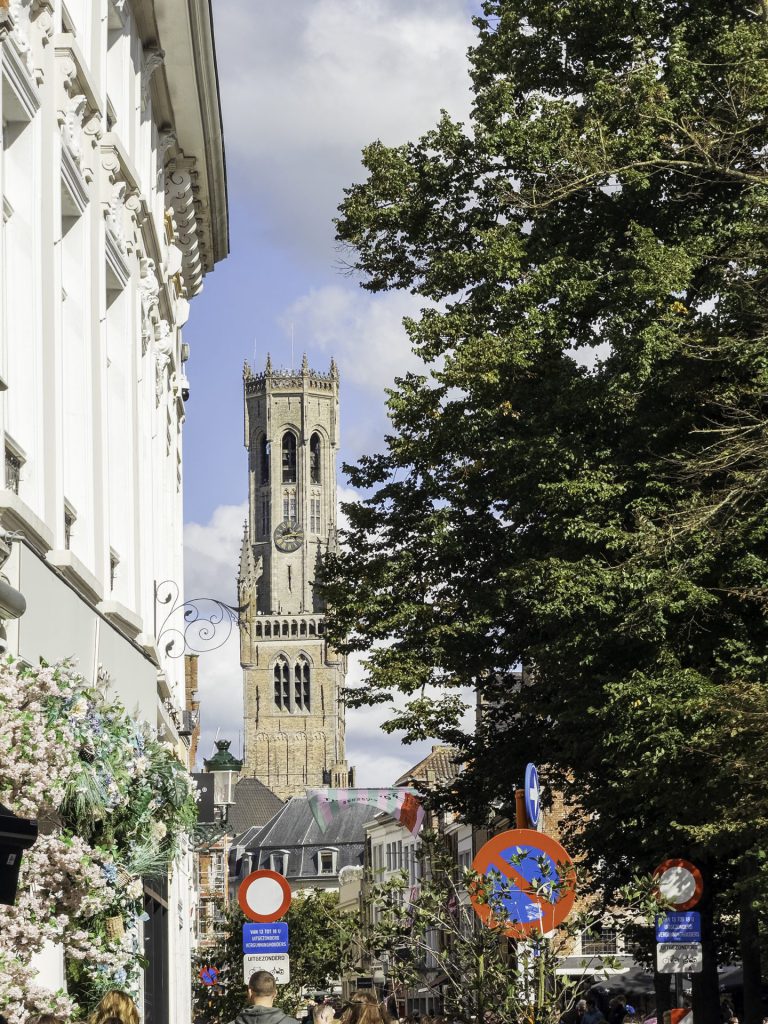 The picture shows a lively street in Bruges with the Belfry, a medieval clock tower, in the background. On the left, we see a facade with white ornaments and floral decoration. On the right, green trees dominate the image. In the foreground: street signs, colourful flags and people walking. This is not a "timeless photo".