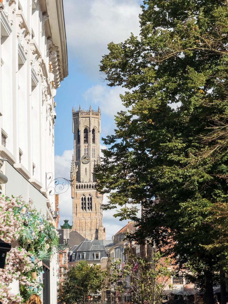 The Belfry tower rises in the centre of the composition, framed by a white-plastered facade with floral decoration on the left and a green tree on the right. In the foreground, there is less disruption: no traffic signs, less crowds - making the tower visually stronger. This composition exudes tranquillity. The vertical lines of the buildings lead the eye upwards, to the crown of the tower that detaches nicely from the sky. The combination of blossom, baroque façade details and historic stone creates a story of transience as well as continuity.