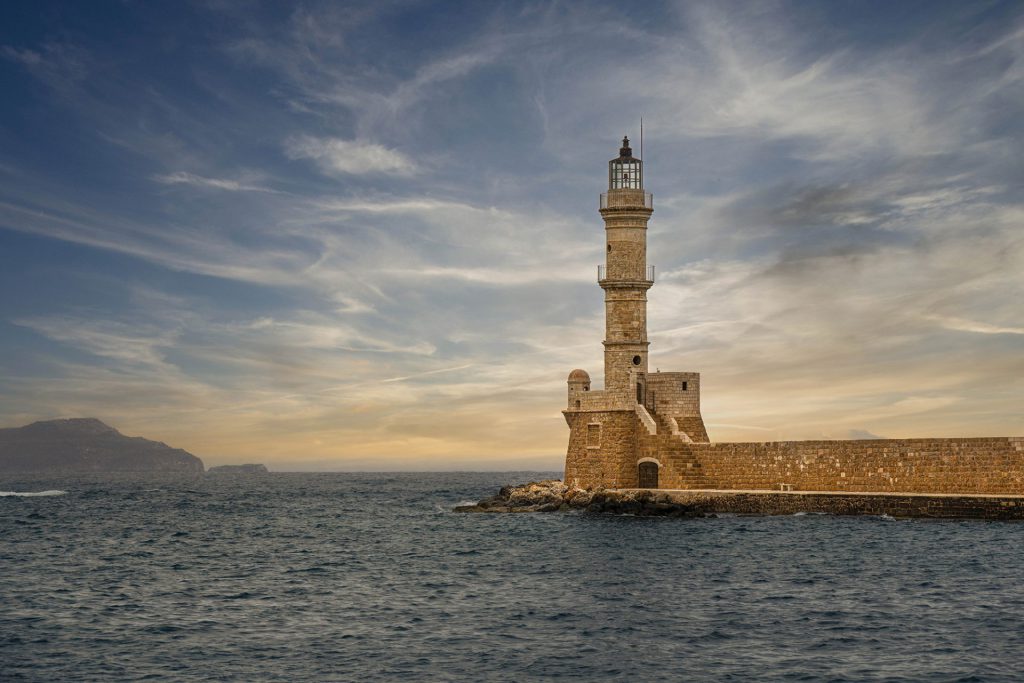 The picture shows the Chania Lighthouse in Crete, Greece - a Venetian lighthouse with iconic architecture on a pier surrounded by calm, dark blue sea water. The sky is impressively dramatic with clouds floating elegantly in a dusky atmosphere. The scene exudes calm, steadiness and a touch of melancholy. The style is realistic with fine art touches thanks to the use of colour and the sky composition. The lighthouse is given an almost symbolic role here as a beacon, both literally and figuratively.