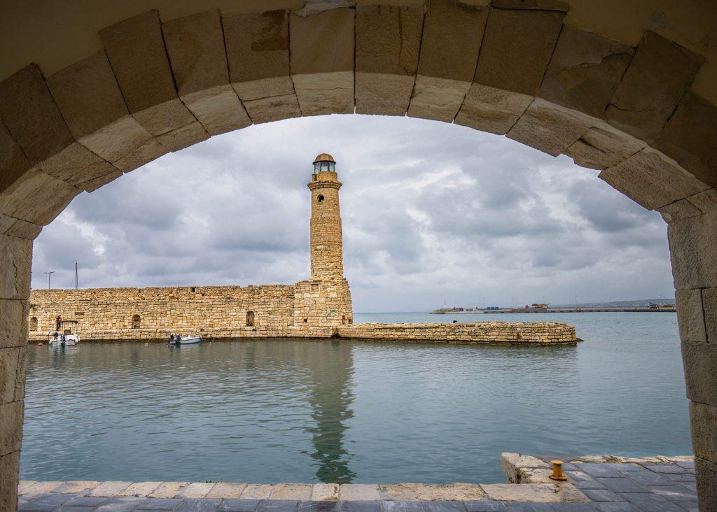 The photo shows the Chania Lighthouse - a Venetian lighthouse with iconic architecture on a pier surrounded by calm, dark blue sea water. The sky is impressively dramatic with clouds floating elegantly in a twilight atmosphere. The scene exudes tranquillity, steadiness and a touch of melancholy. Thanks to the use of colour and the composition of the sky, the style is realistic, with touches of fine art. The lighthouse is given an almost symbolic role here as a beacon, both literally and figuratively. (© Guy Lambrechts) photo tour Crete