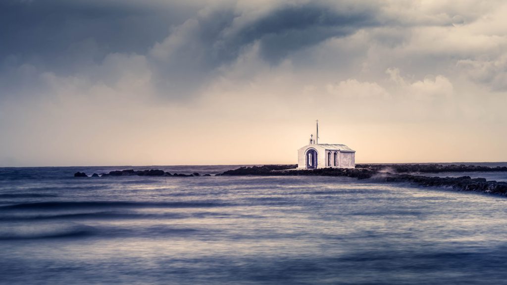 A small, white chapel of Agios Nikolaos in Georgioupoli, stands on a narrow, rocky path leading into the sea. Threatening clouds in deep blue and grey fill the sky. The light falls from behind and frames the chapel with a soft glow. The scene exudes melancholy and a touch of mysticism. The style leans towards fine art landscape photography, focusing on atmosphere and minimalism. The picture exudes calm, isolation and contemplation. The chapel symbolises hope and spirituality in a turbulent world. The photo tells of isolation and devotion - a path to inner peace. This photo with a shutter speed of 25 seconds was taken during a rainstorm, using a 10-stop Big Stopper filter. In post-processing, rain splatters were carefully removed from the lens.