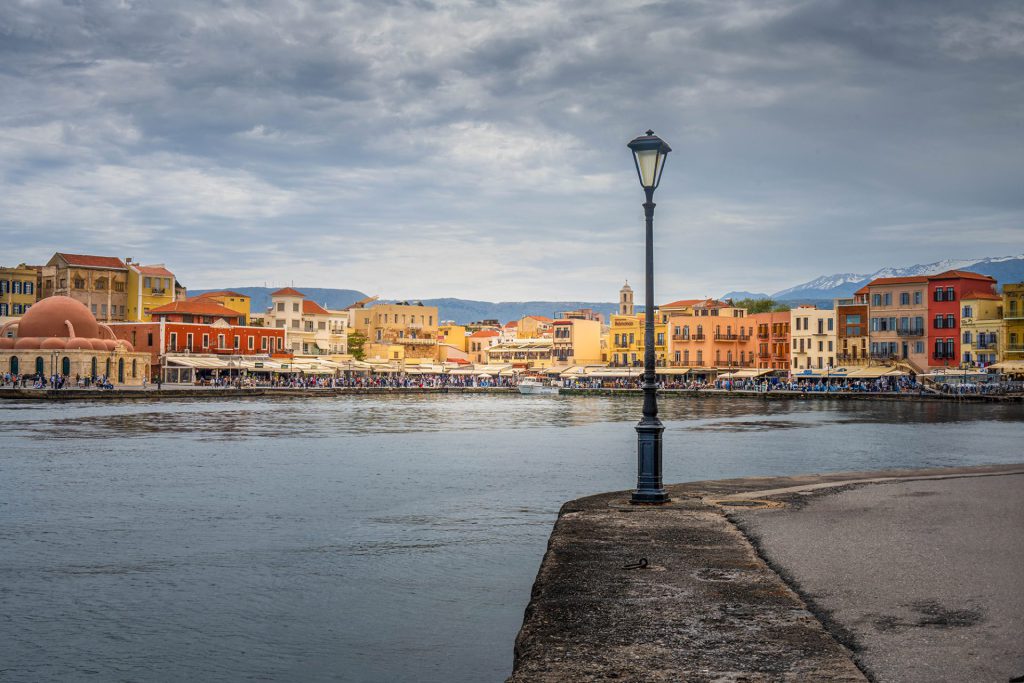 The picture shows a colourful port city of Chania inb Crete under an overcast sky. At the centre is a lamppost at the edge of a quay. In the background, we see pastel-coloured buildings, an old mosque with a dome, and snow-capped mountains in the distance. The water is calm, and there are many people along the promenade. Artful review: The picture balances perfectly between urban vibrancy and natural tranquillity. The colourful facades contrast nicely with the austere sky and calm water. The composition emphasises isolation and focus, especially with the central placement of the lamppost. This acts as a visual anchor and introduces a certain stillness against the bustle in the background.