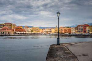 The picture shows a colourful port city under an overcast sky. At the centre is a lamppost at the edge of a quay. In the background, we see pastel-coloured buildings, an old mosque with a dome, and snow-capped mountains in the distance. The water is calm, and there are many people along the promenade. Artful review: The picture balances perfectly between urban vibrancy and natural tranquillity. The colourful facades contrast nicely with the austere sky and calm water. The composition emphasises isolation and focus, especially with the central placement of the lamppost. This acts as a visual anchor and introduces a certain stillness against the bustle in the background.