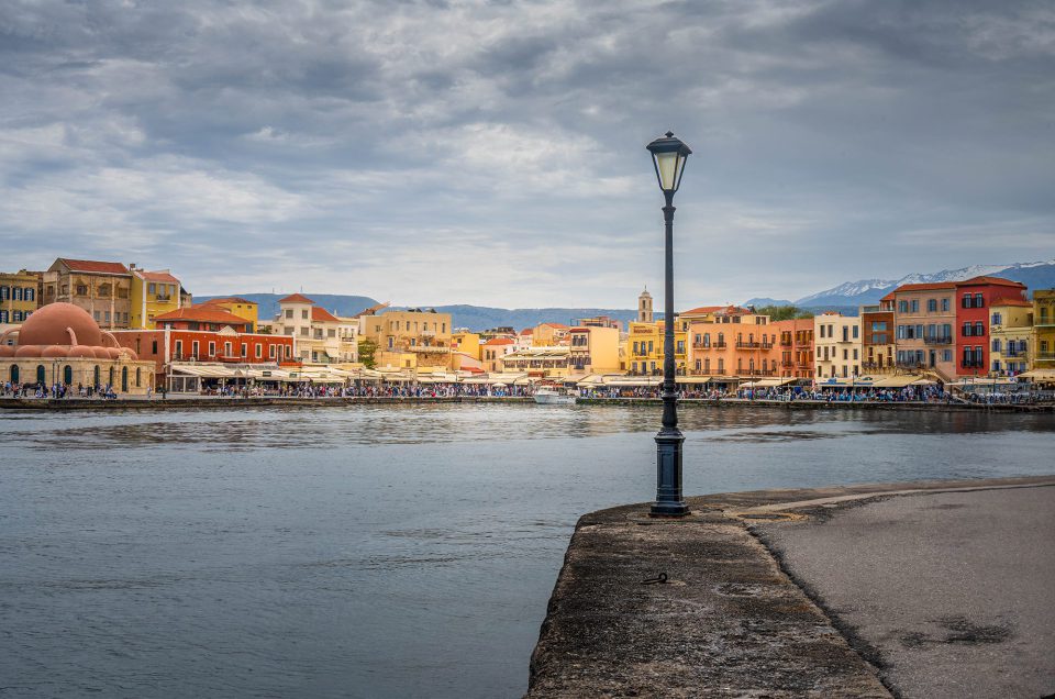 The picture shows a colourful port city under an overcast sky. At the centre is a lamppost at the edge of a quay. In the background, we see pastel-coloured buildings, an old mosque with a dome, and snow-capped mountains in the distance. The water is calm, and there are many people along the promenade. Artful review: The picture balances perfectly between urban vibrancy and natural tranquillity. The colourful facades contrast nicely with the austere sky and calm water. The composition emphasises isolation and focus, especially with the central placement of the lamppost. This acts as a visual anchor and introduces a certain stillness against the bustle in the background.